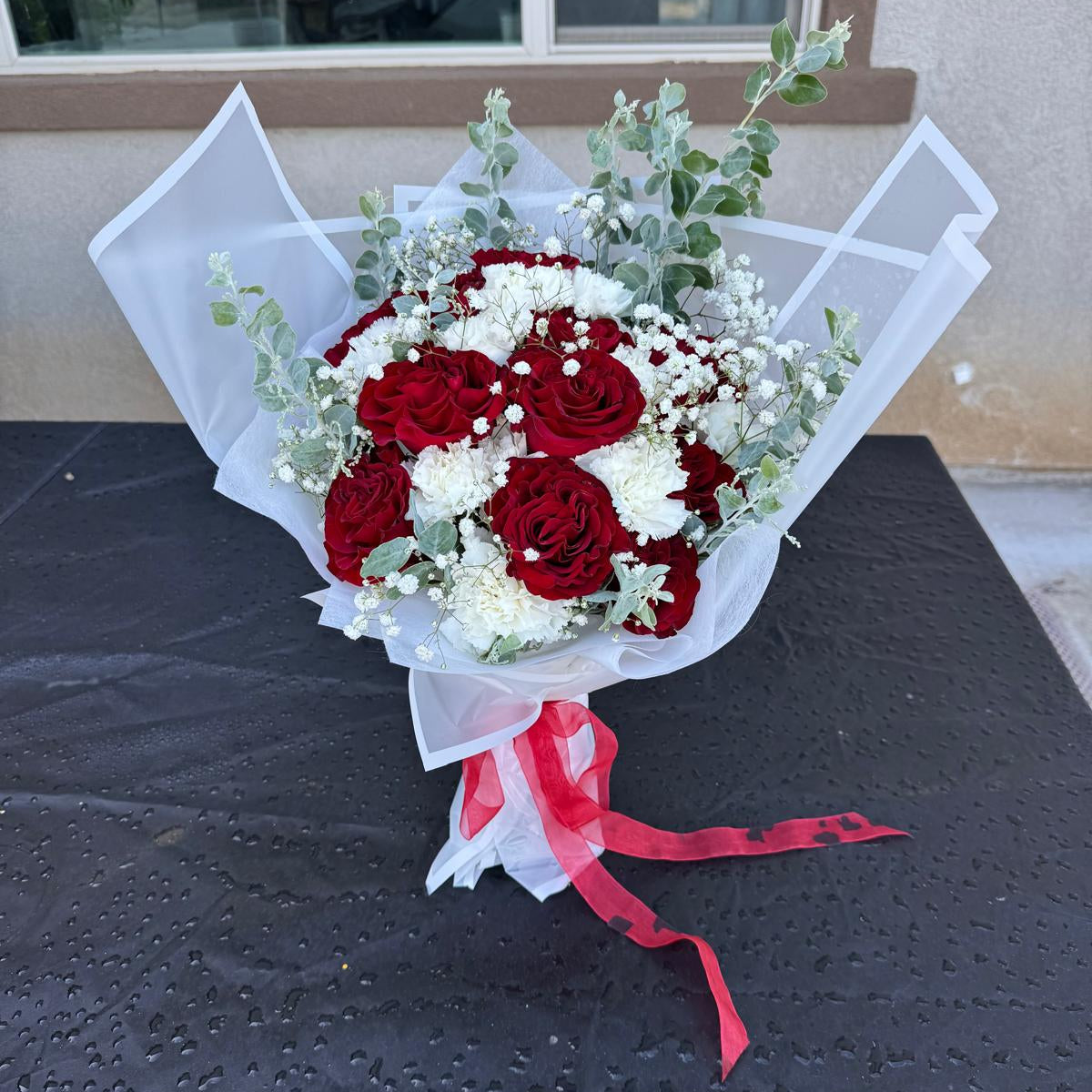 Signature Veterans Day bouquet with red roses, white carnations and baby’s breath wrapped in white paper with red ribbon by Nai Pascoal Bouquets.