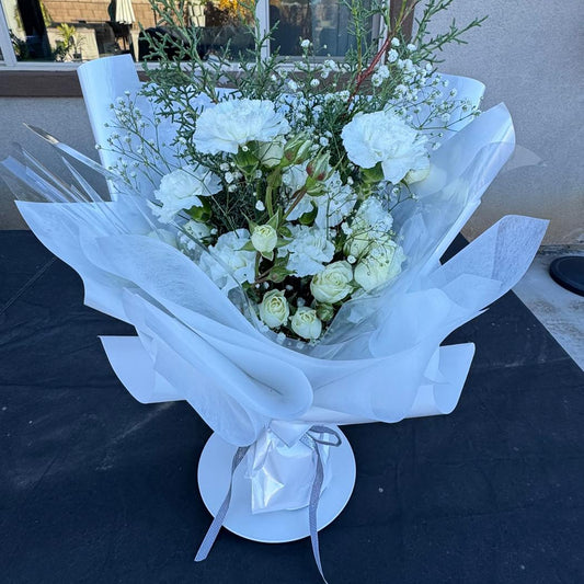 A hand-tied white winter bouquet with carnations, spray roses, and delicate winter greenery, wrapped in layered white paper – Nai Pascoal Bouquets Winter Whisper.