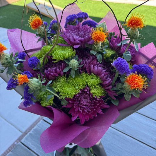 Witch’s Brew Bouquet featuring purple, orange, and green flowers with curly willow branches, wrapped in black.