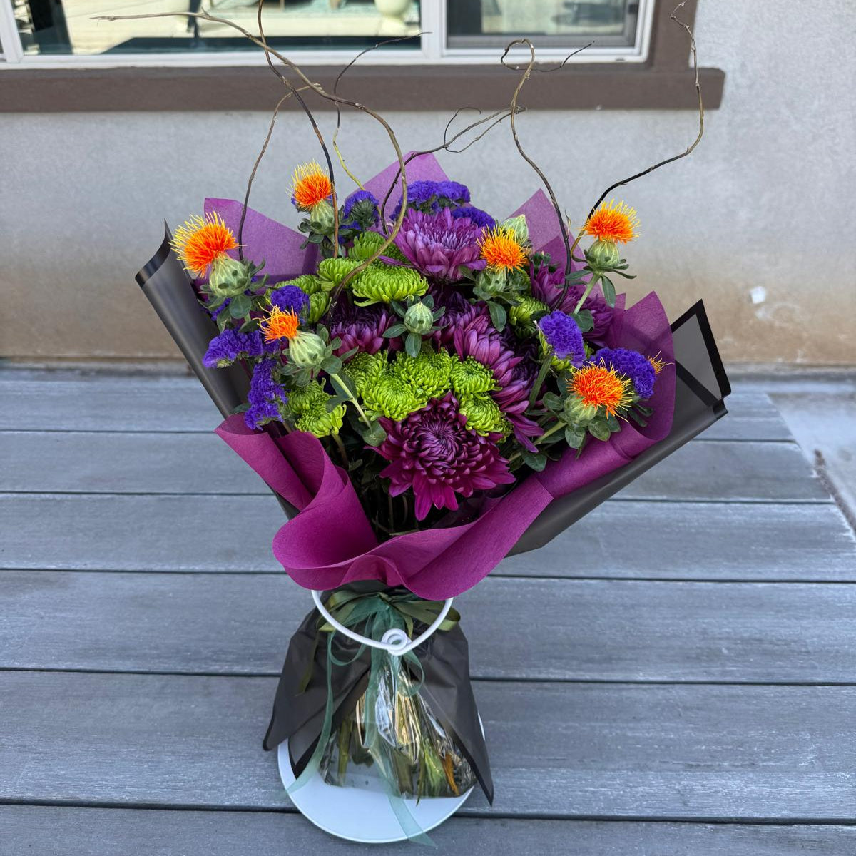 Witch’s Brew Bouquet featuring purple, orange, and green flowers with curly willow branches, wrapped in black.